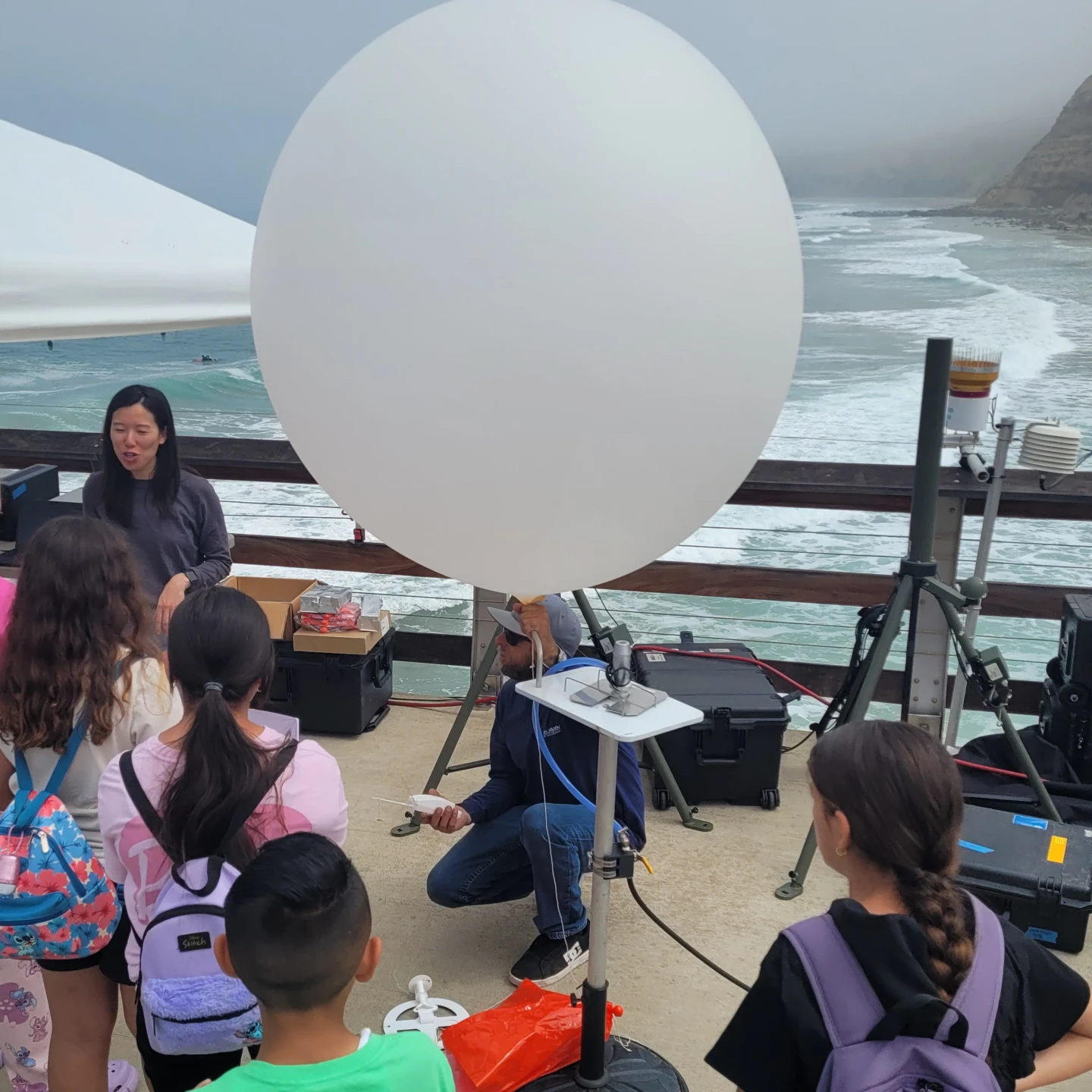 Someone showing a large white weather balloon to a group of children on a coastal pier.