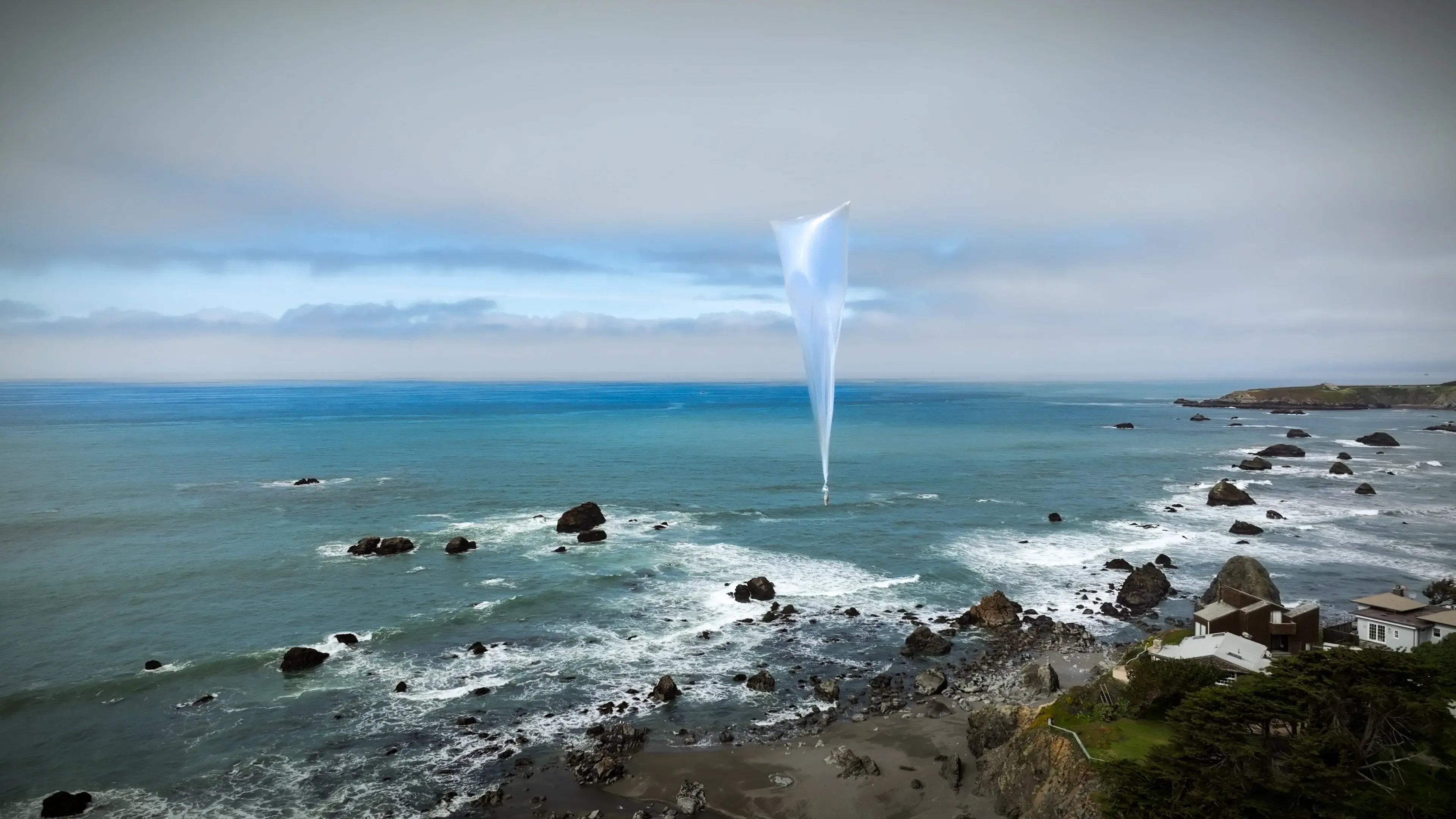 A white weather balloon above a rocky shoreline and blue ocean beneath low clouds.