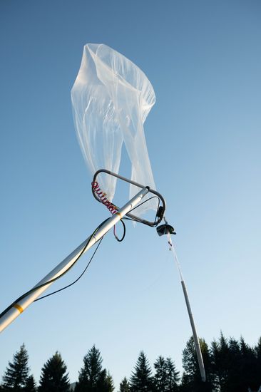 A partly inflated clear balloon lifting off the launch rig's arm against a blue sky.