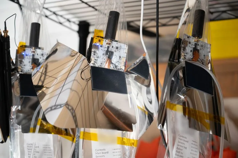 Balloon sensor packages, with circuit boards, solar panels and reflective foil, hanging on a rack.